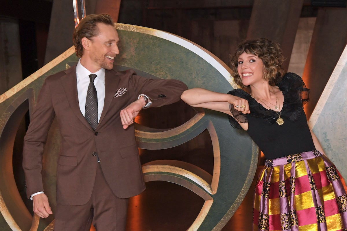 'Loki' stars Tom Hiddleston and Sophia Di Martino bump elbows while standing in front of a display of the show's logo