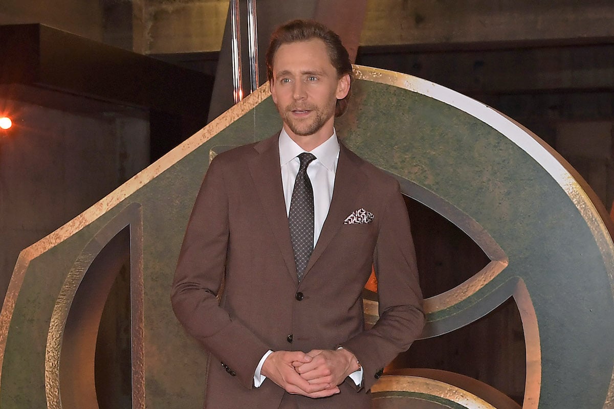 Tom Hiddleston wears a suit and poses in front of a display of the 'Loki' logo