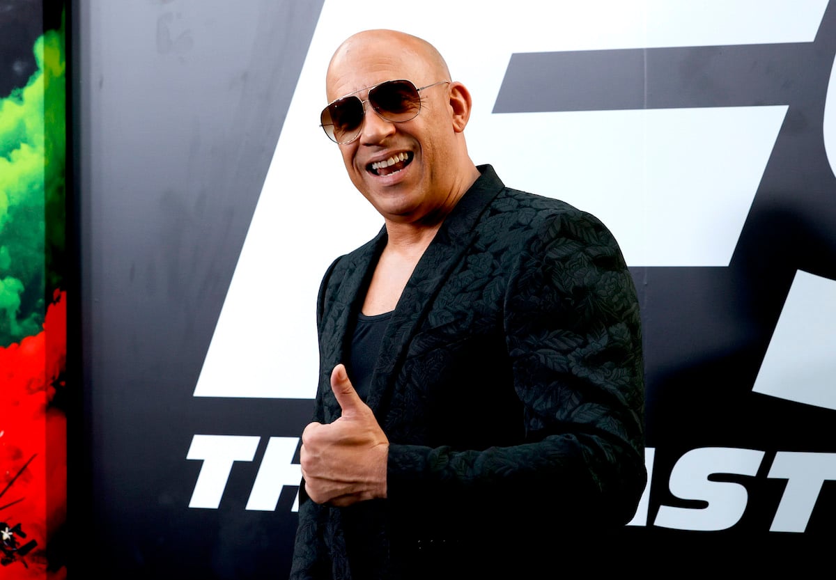 Vin Diesel wears black, smiles, and offers a thumbs up while standing in front of art for ‘F9: The Fast Saga’