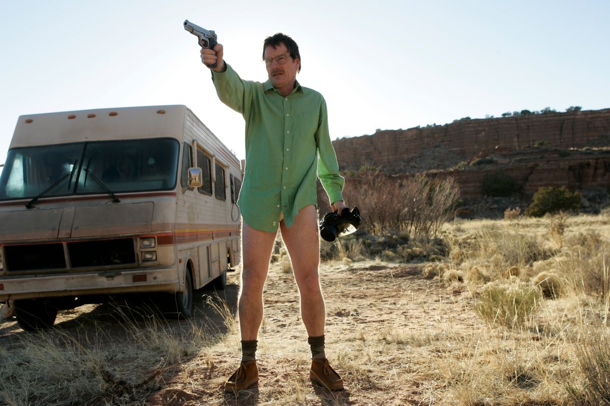 Walter White points a gun as he stands in his underwear, shoes, and a green button-up shirt.