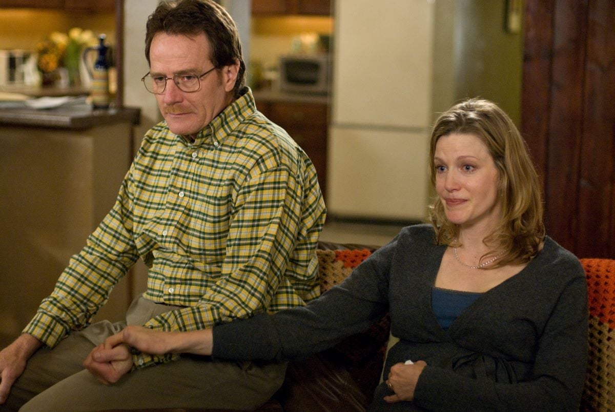 Bryan Cranston as Walter and Anna Gunn as Skyler White hold hands while sitting on a couch together.