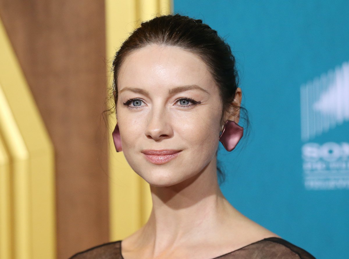 Caitriona Balfe with her hair slicked back in a low bun and wearing a black gown with a black mesh neckline at the 'Outlander' Season 5 premiere. She stands in front of a teal background