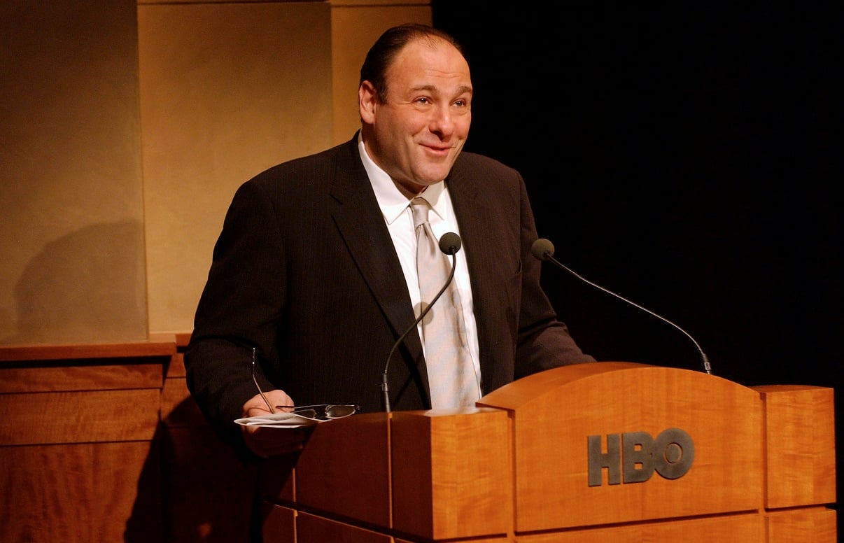 James Gandolfini smiles as he stands at a podium with the HBO logo on it.