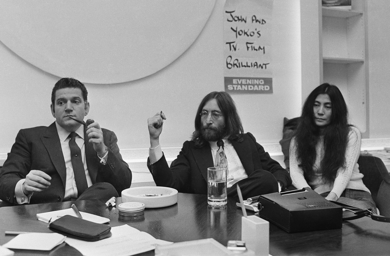 John Lennon, wearing a suit, sits at a table between Allen Klein and Yoko Ono, 1969.