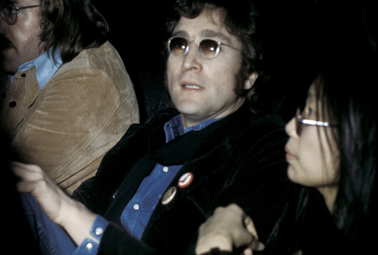 John Lennon stares at the camera while sitting in a theater with May Pang, 1974.