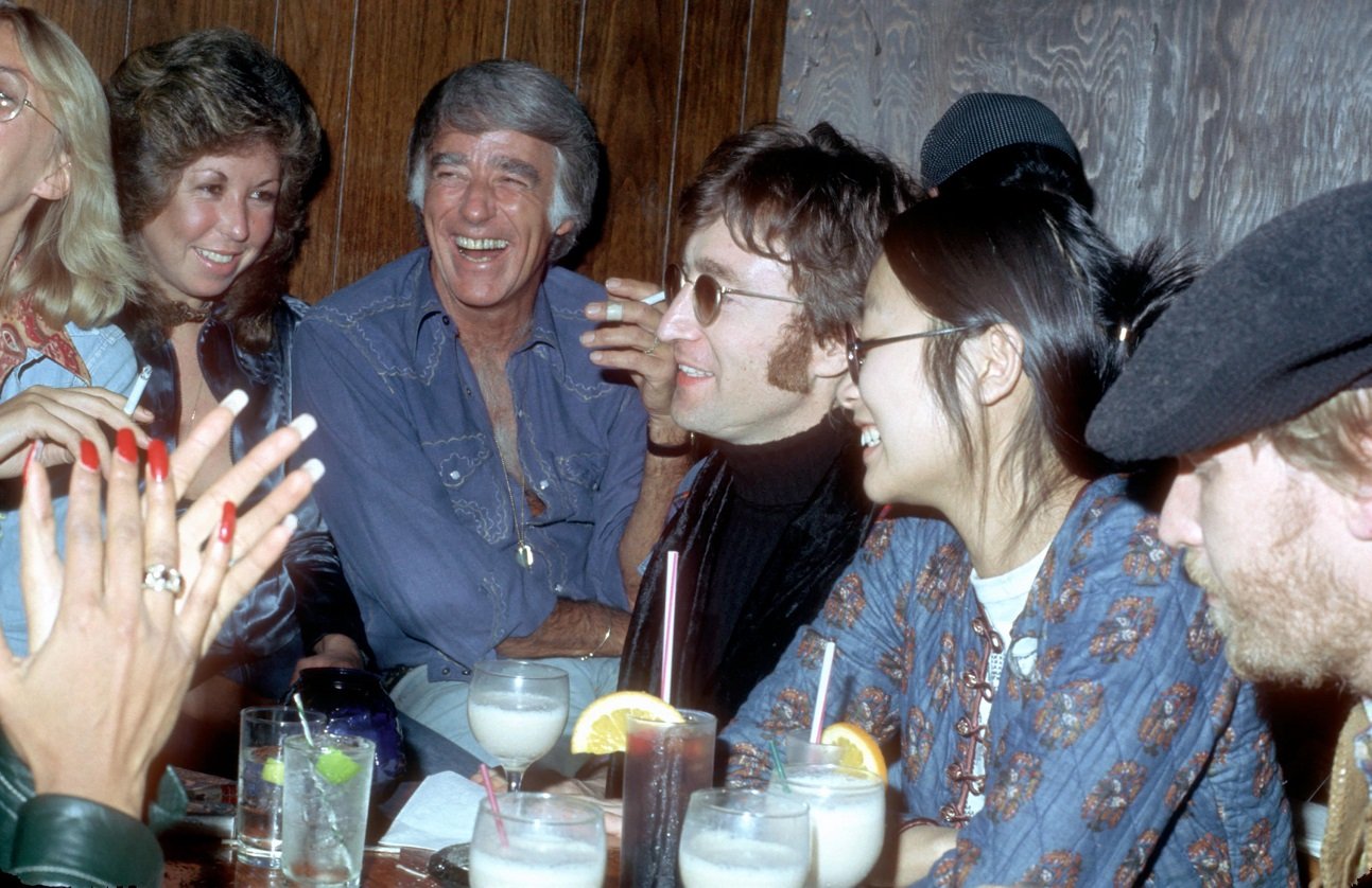 John Lennon, May Pang, Harry Nilsson, and friends pictured seated together at a club, laughing, with cocktails in front of them