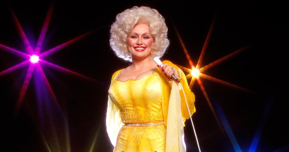 Dolly Parton is photographed in front of a black background with rainbow lights. She's in a yellow dress and holding a white microphone.