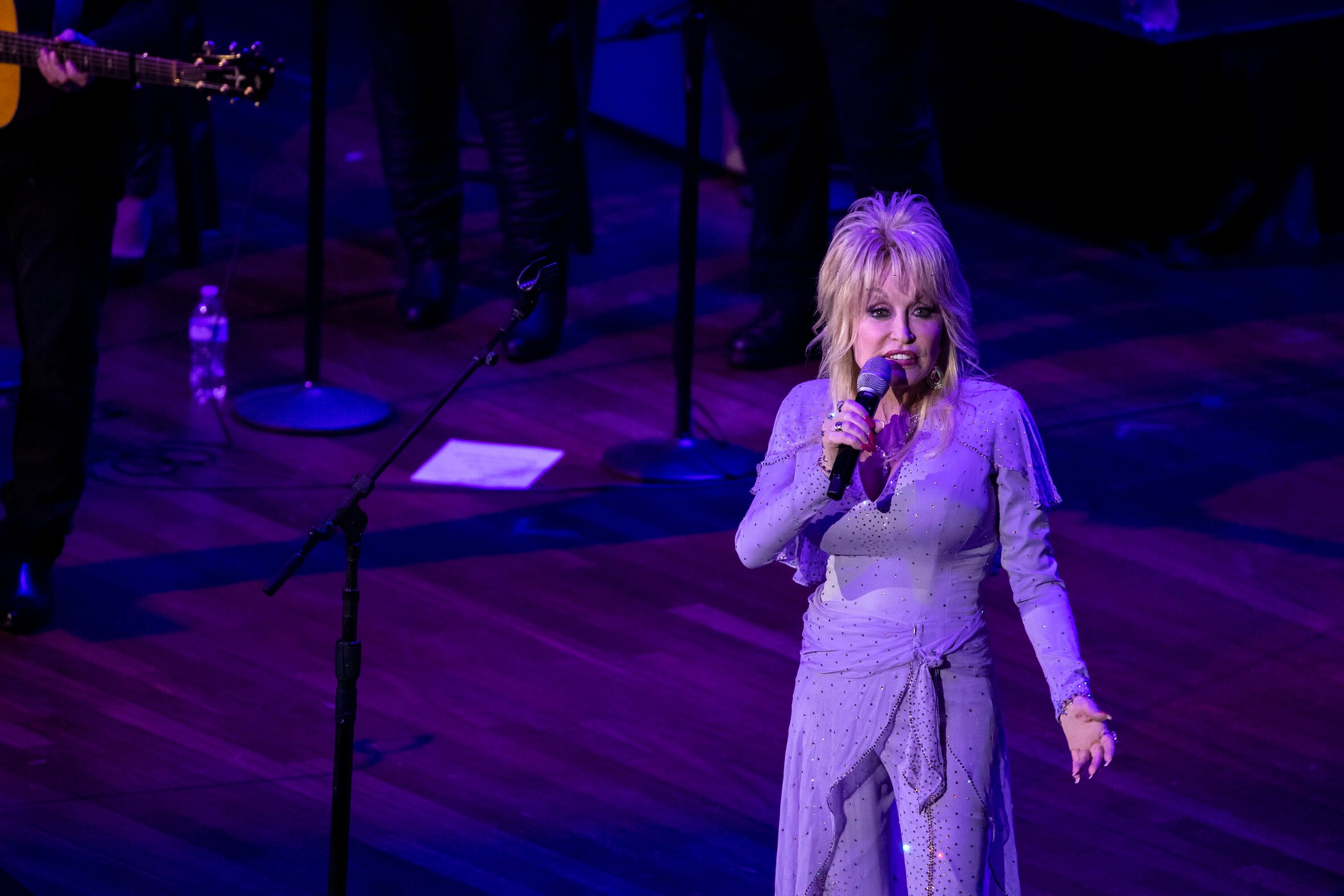 Dolly Parton performs on stage at The Gift of Music Concert. She's singing into a microphone in an all-purple outfit.