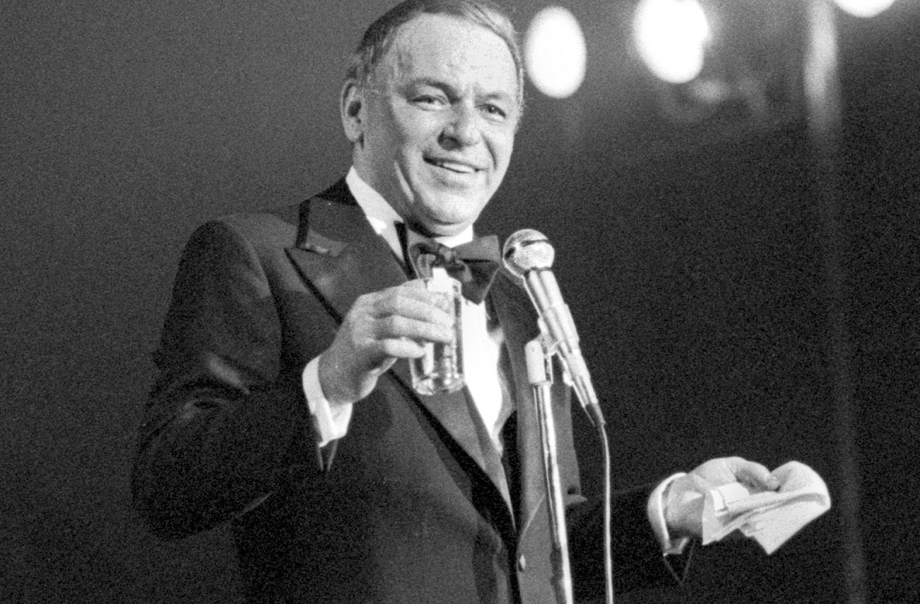 Frank Sinatra smiles on stage while holding a glass in one hand and a handkerchief in the other, 1975.