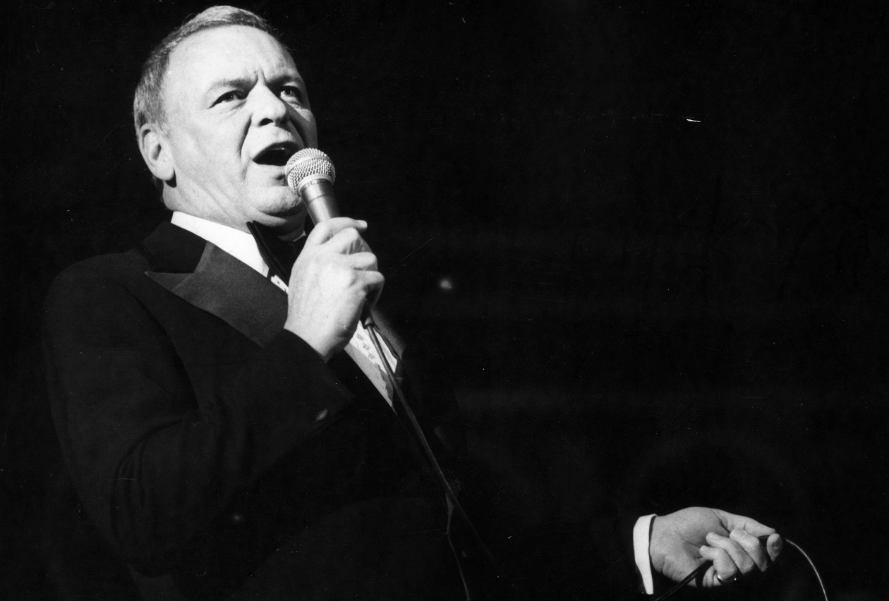 Closeup of Sinatra from the waist up holding a microphone and singing into it in 1976