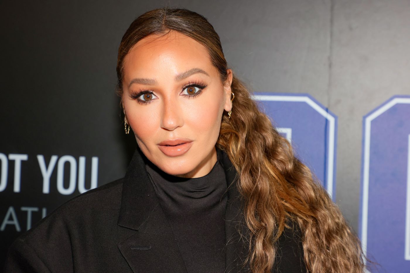Adrienne Bailon stands in front of a black backdrop with a black turtleneck.