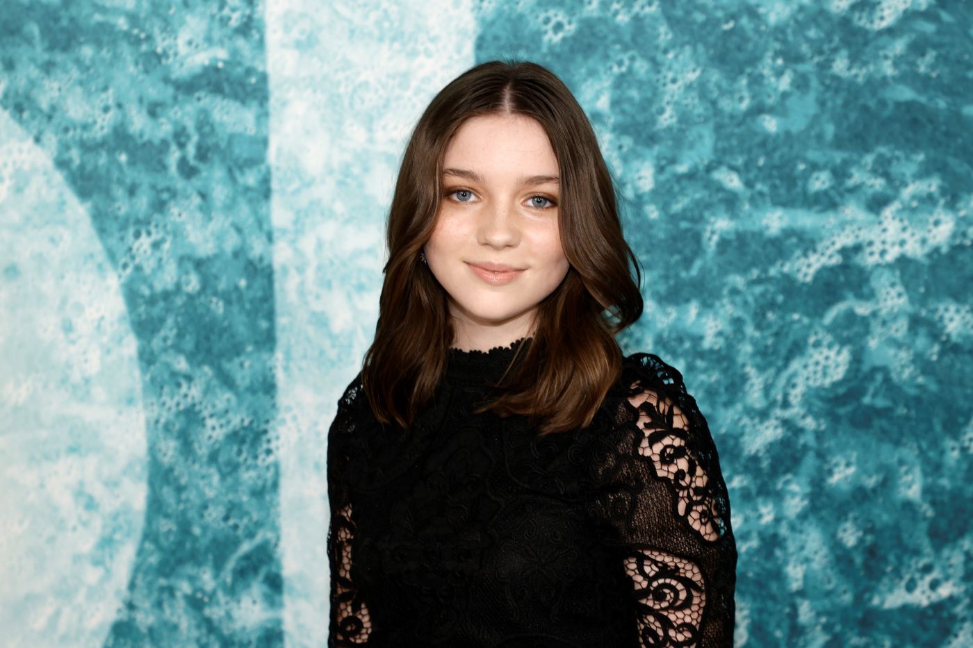 Alexa Swinton is in-front of a ocean colored and textured background wearing a black blouse with with black full-length lace sleeves.