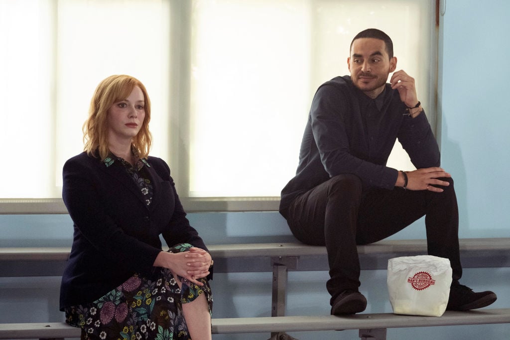 Christina Hendricks as Beth Boland and Manny Montana as Rio sit on the bleachers together. Rio looks over at Beth slyly.