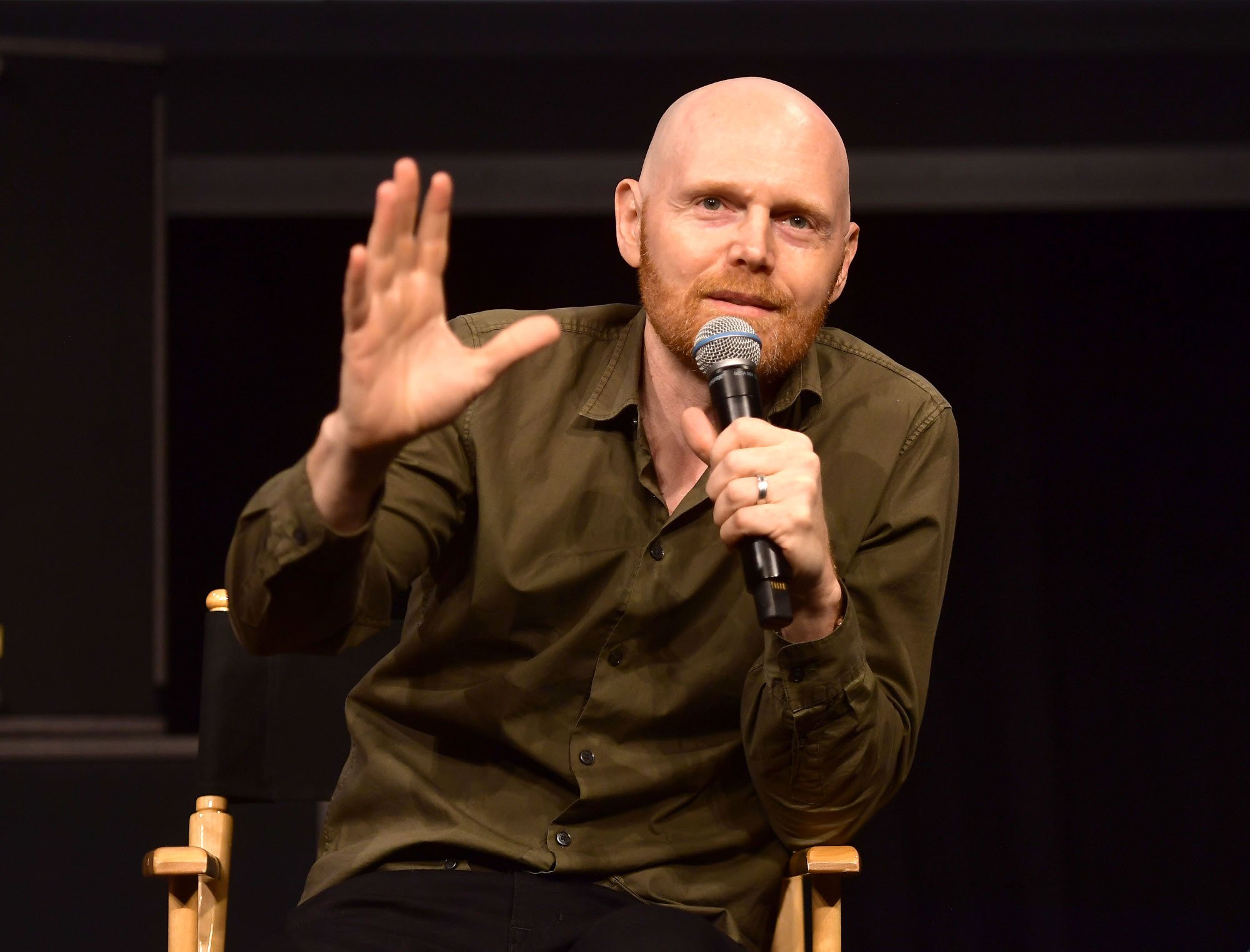 Bill Burr wearing a brown shirt, holding one hand up, and speaking into a microphone