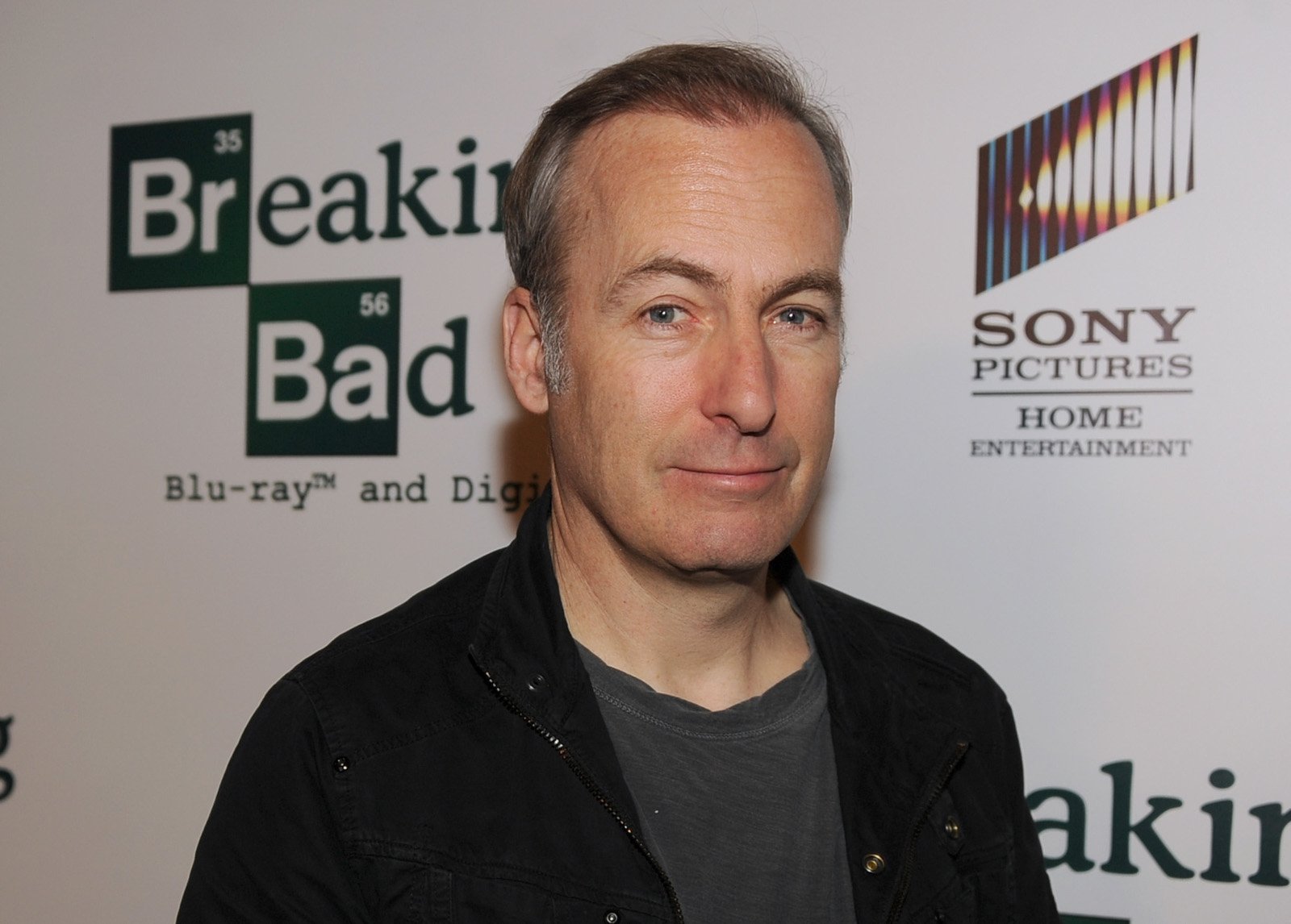 'Better Call Saul' star Bob Odenkirk stands in front of a 'Breaking Bad' and AMC wall wearing a grey shirt and black jacket