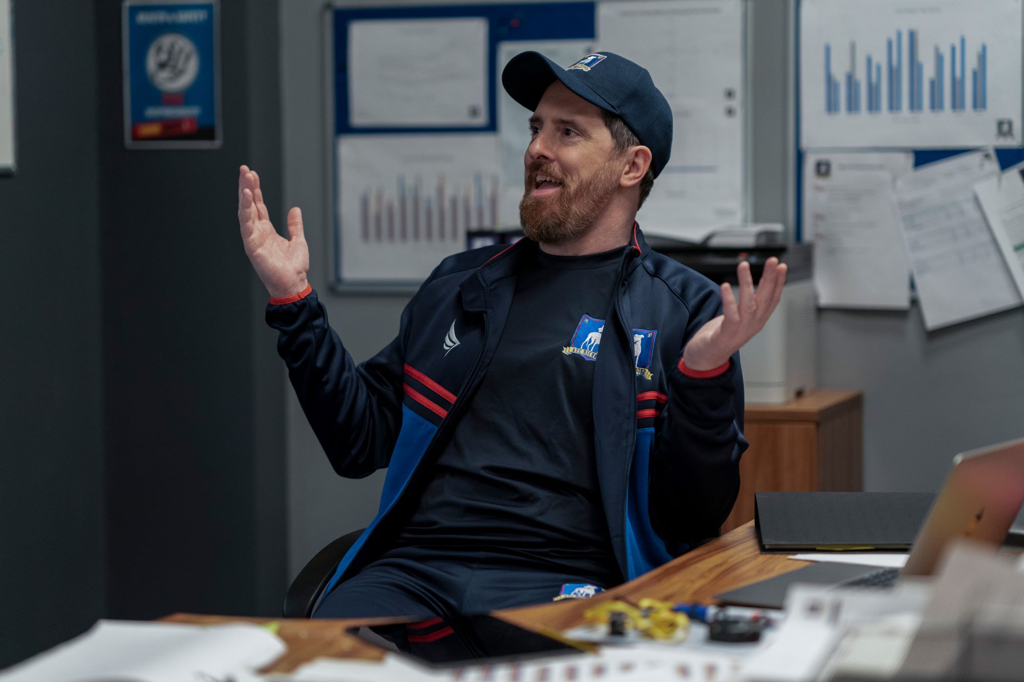 A scene from 'Ted Lasso' with Brendan Hunt's character sitting in a chair in the coach's office with hands thrown up wearing a black shirt with a blue vest with red stripes on the chest and a blue hat. 