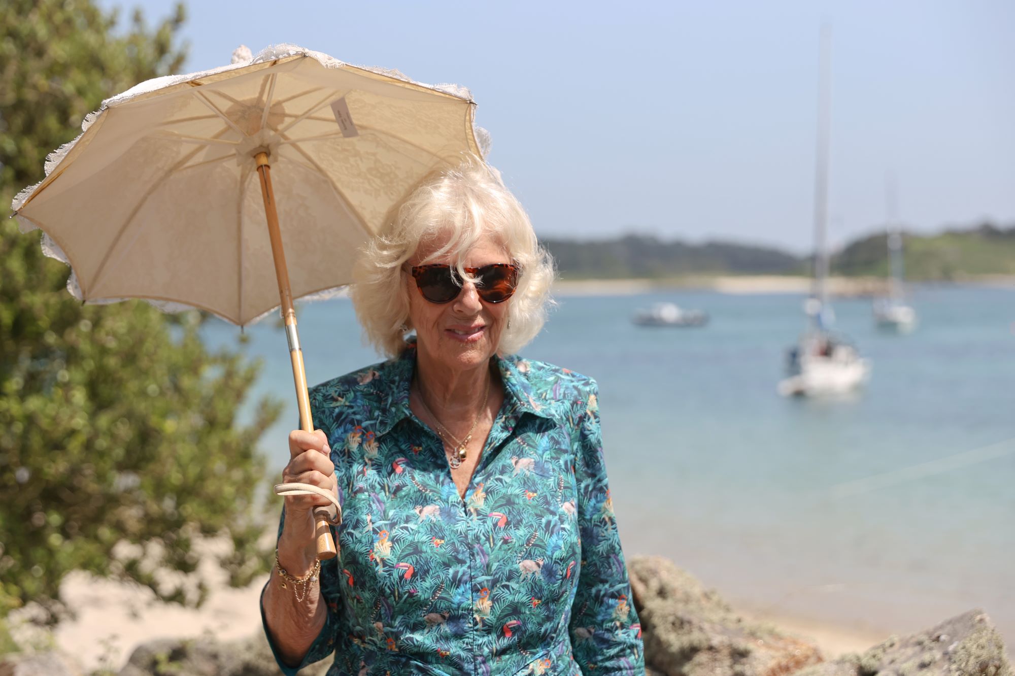 Camilla parker Bowles is walking along a waterfront with boats on it holding a white umbrella dressed in a tropical green, red, and yellow shirt.