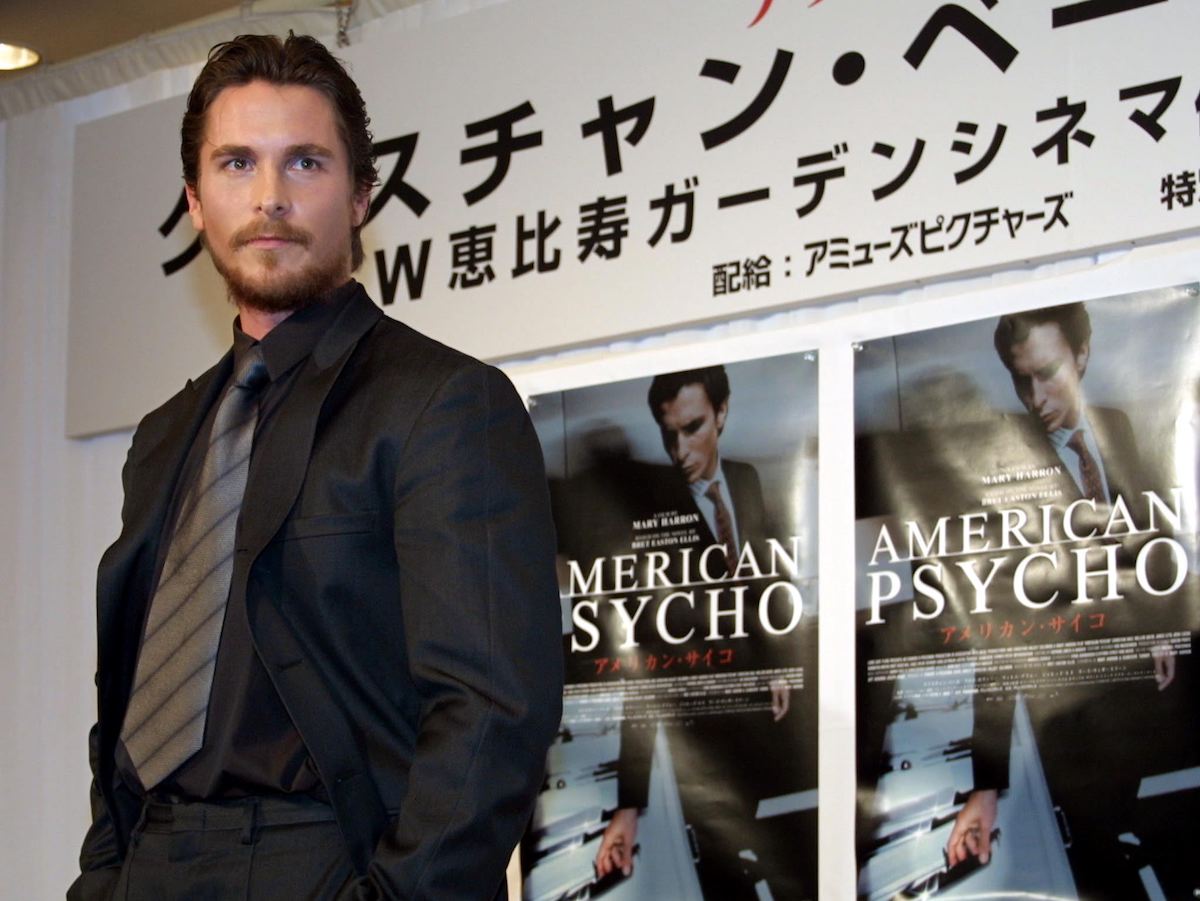 Christian Bale wears a suit and poses in front of ‘American Psycho’ posters