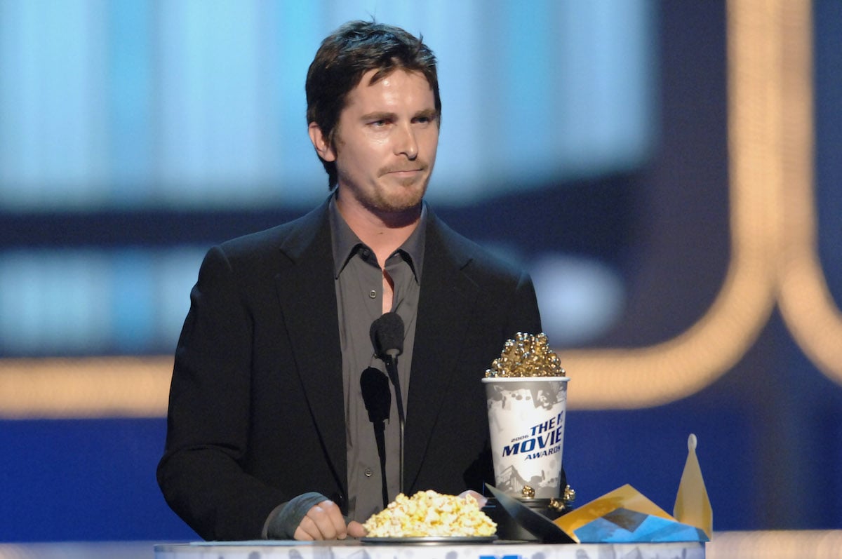 Christian Bale stands at a podium dressed in a jacket