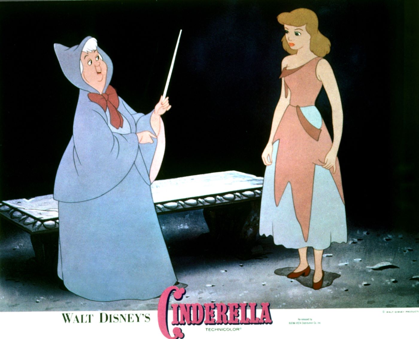 The animated Cinderella in her rags and the fairy godmother in her blue dress with her wand stand in front of a black background with a stone bench behind them.