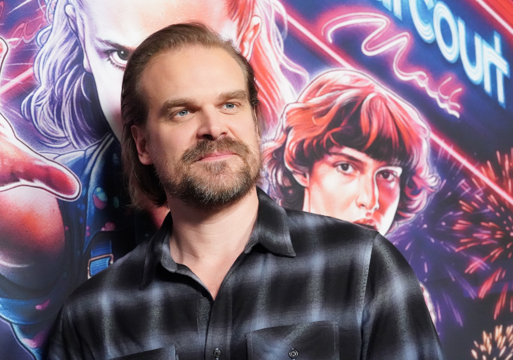 Stranger Things star David Harbour wearing a black and white flannel shirt and standing in front of a Stranger Things wall