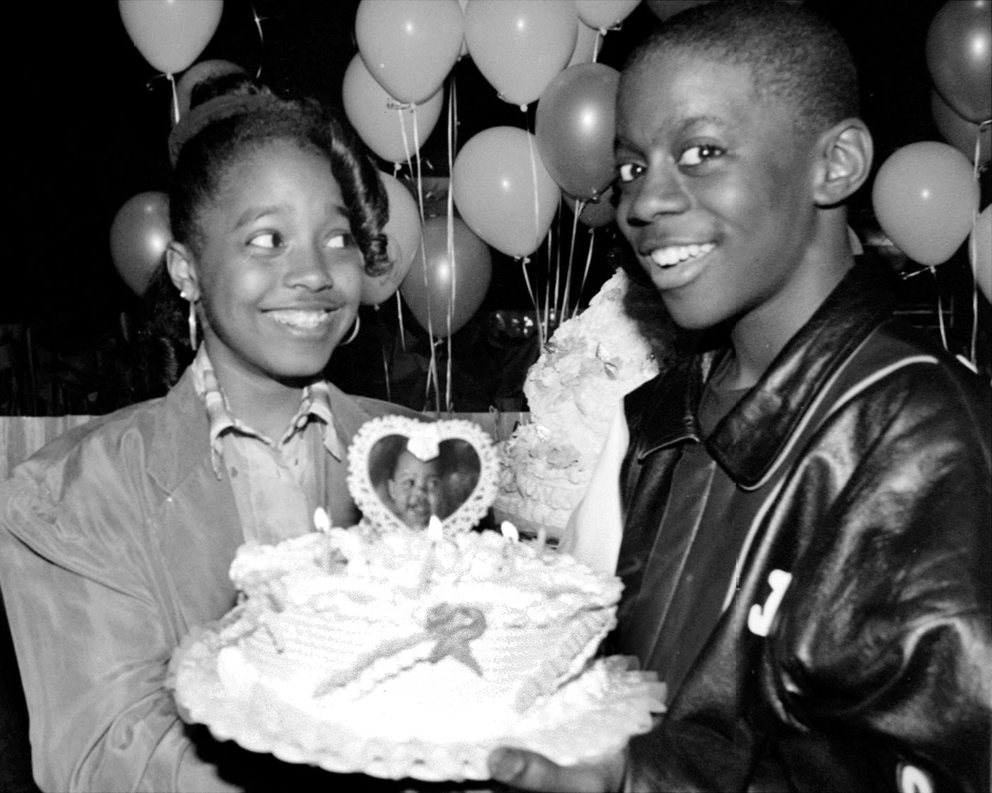 A black and white photo of Keshia Knight Pulliam and Deon Richmond holding a birthday cake with a heart on top and balloons in the background.