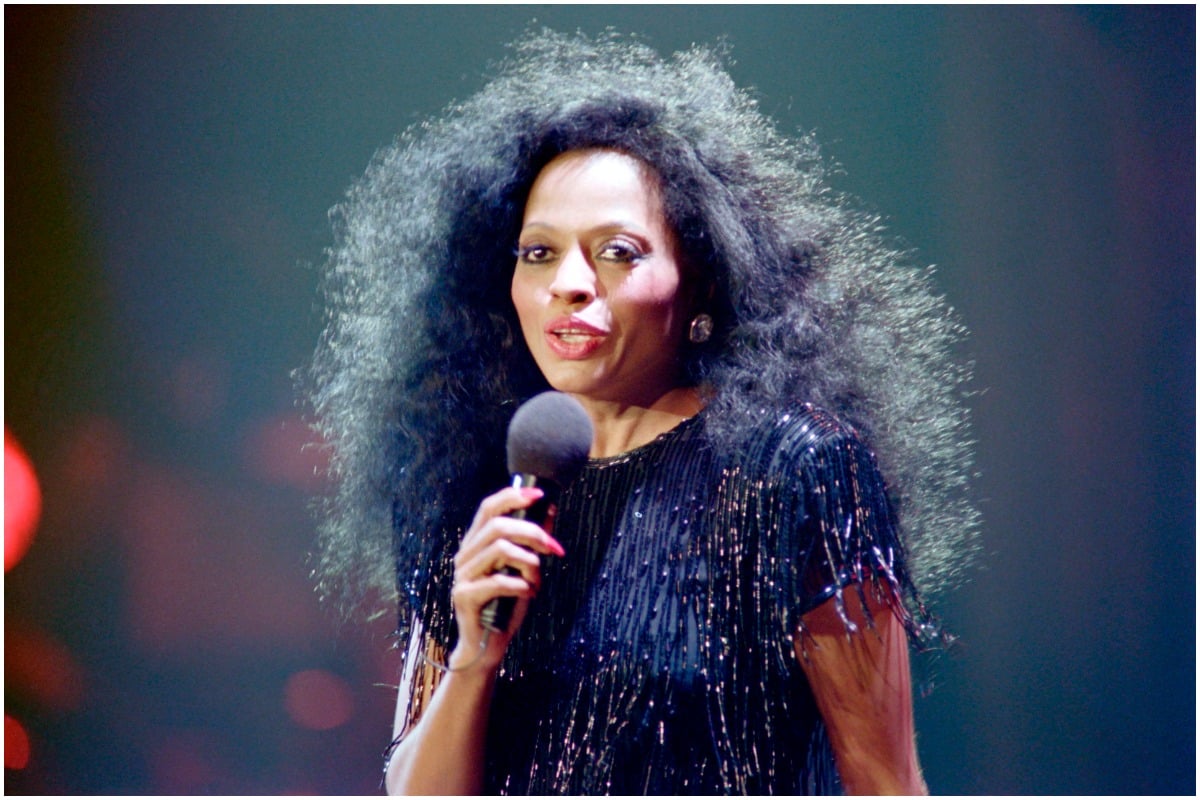 Diana Ross holding a microphone and talking while wearing a black top.