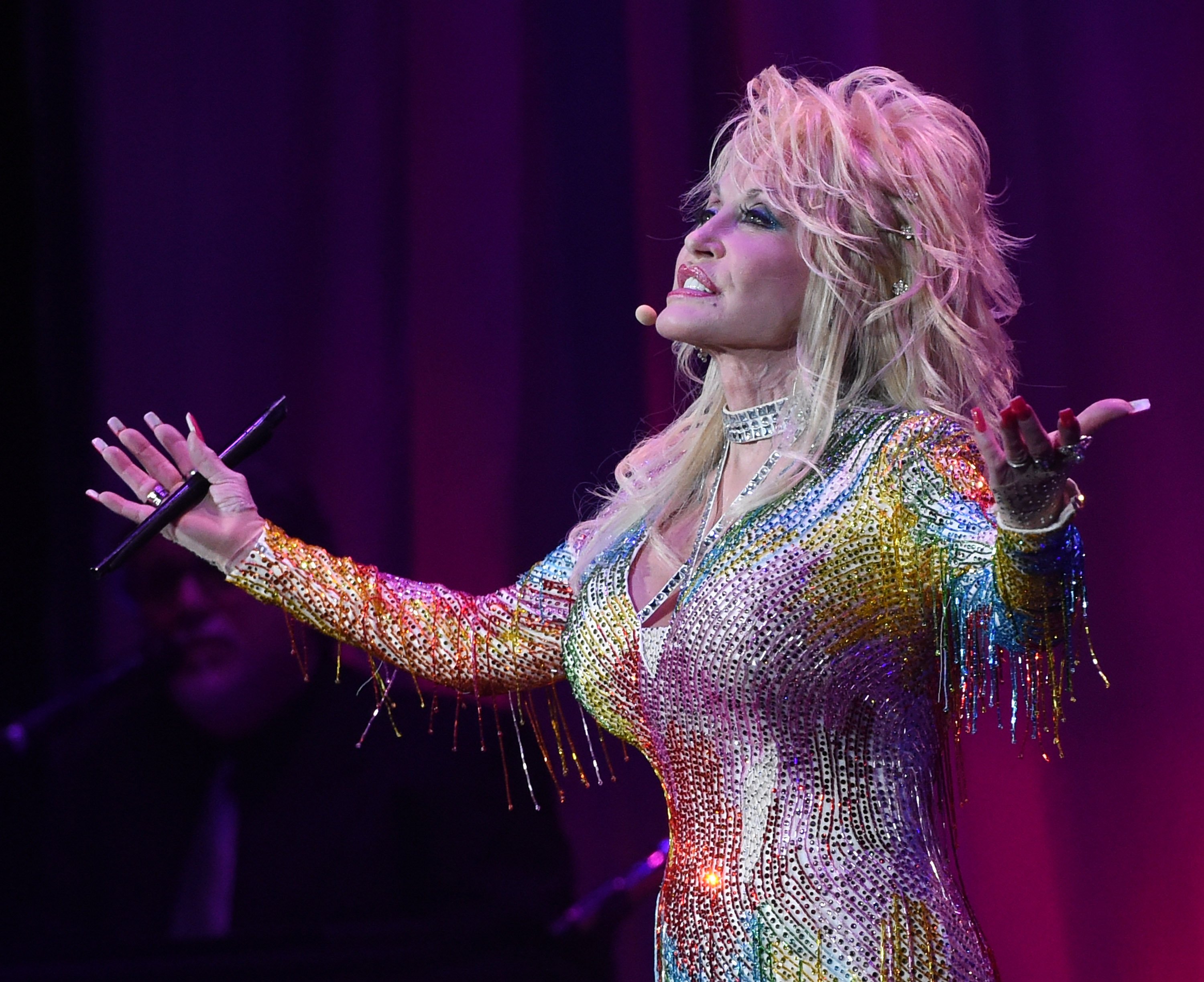 Dolly Parton performs on stage. She's wearing a rainbow, sparkly outfit.