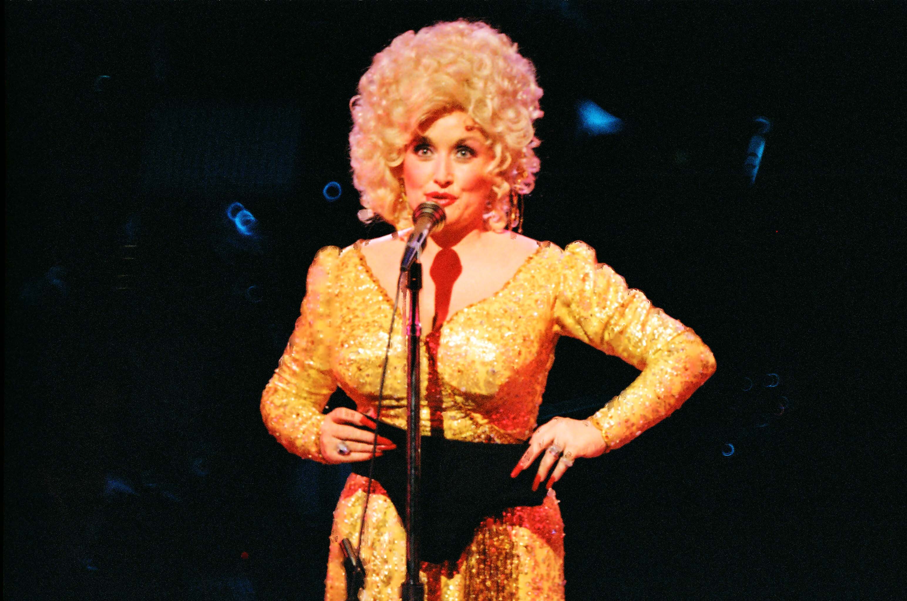 Dolly Parton stands on stage in a gold dress. She's speaking into a microphone and hold black underwear.