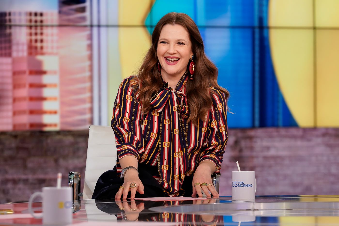 Drew Barrymore sitting in a black and orange stripped shirt in front of a glass desk with a red, blue, and yellow blurred background.