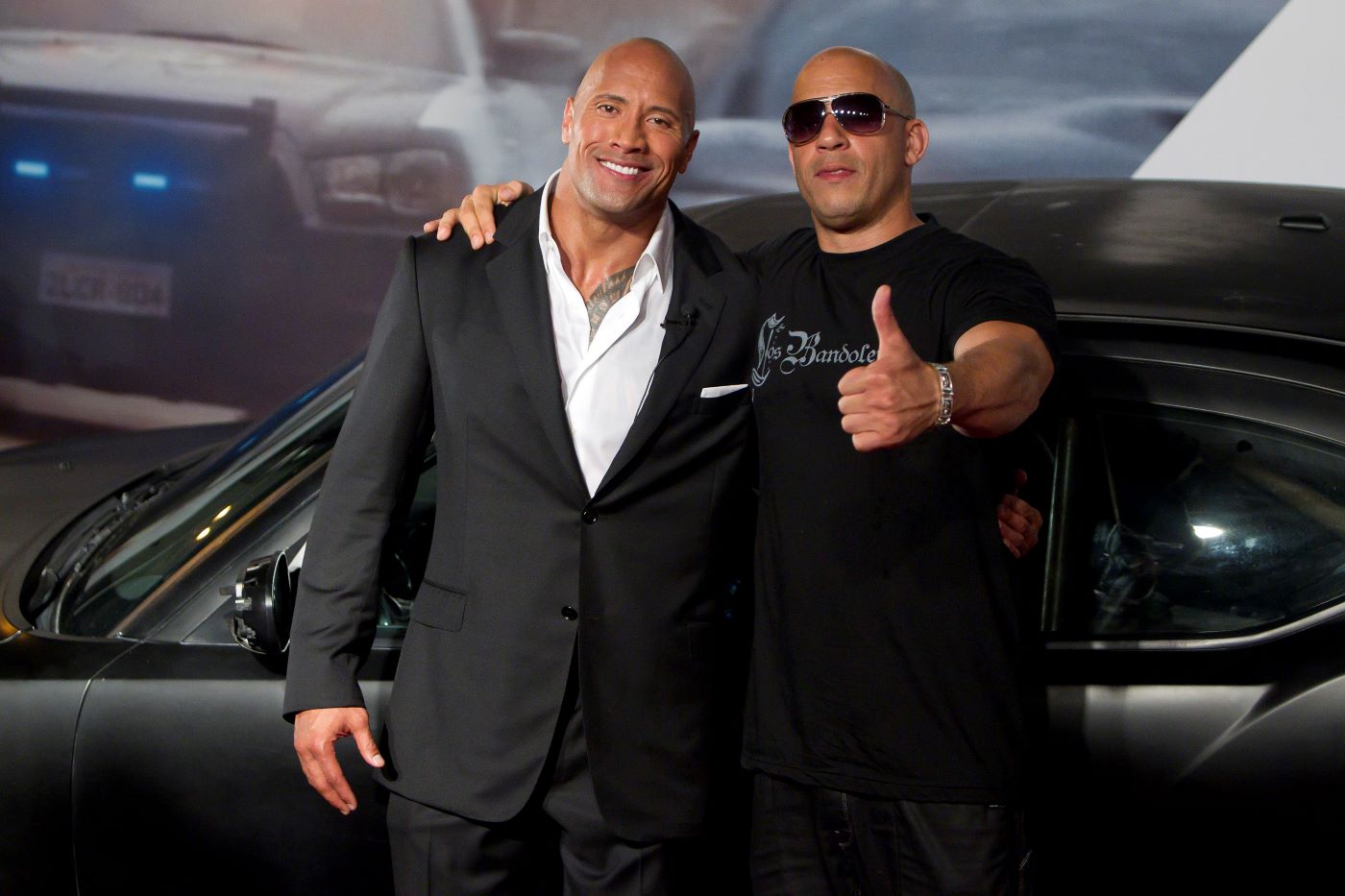 Dwayne Johnson and Vin Diesel from 'The Fast and Furious' movies stand in front of a black sports car. Johnson is wearing a black suit with a white button-up shirt and Diesel is wearing a black shirt with white writing and black jean pants.