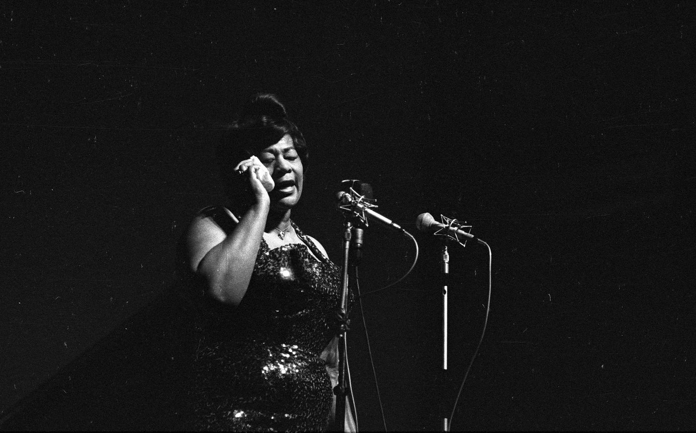A black-and-white photo of Ella Fitzgerald standing as she sings into a microphone on a darkened stage
