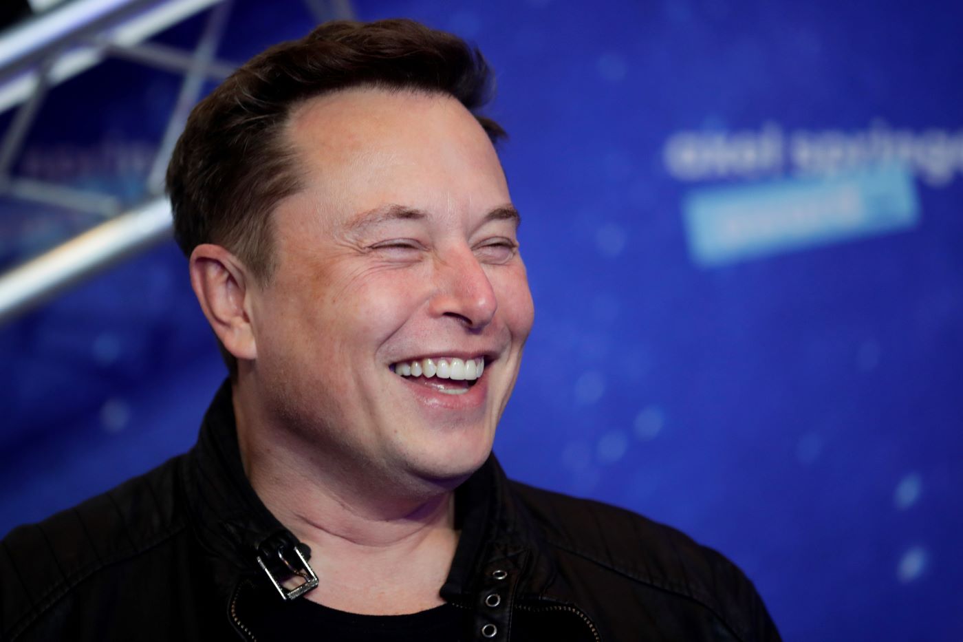 Closeup on Elon Musk's face while smearing wearing a black shirt against a blue background.