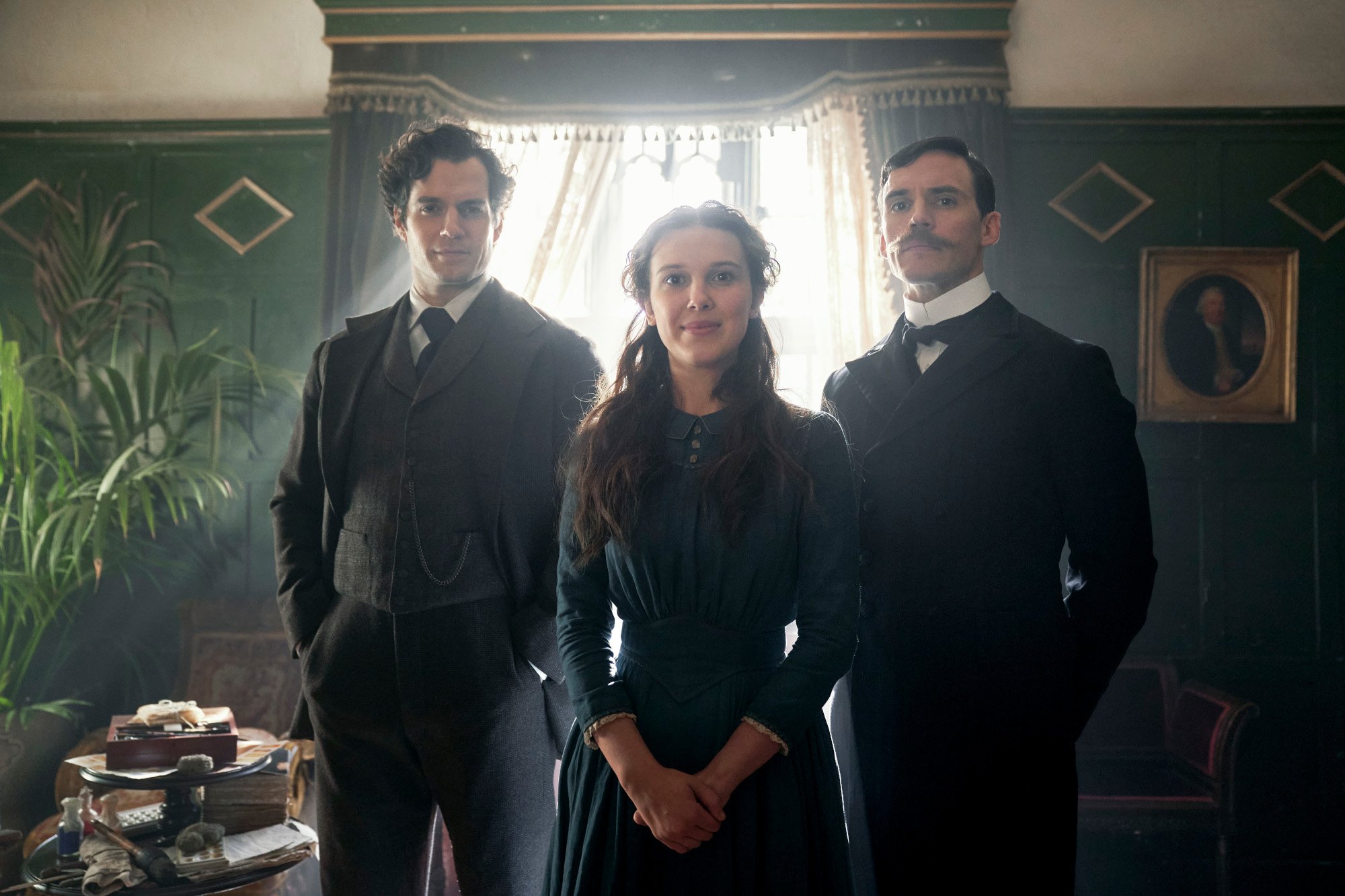 Henry Cavill, Millie Bobby Brown, and Sam Claflin wear dark colors and stand in front of a dark green wall in Netflix's 'Enola Holmes'