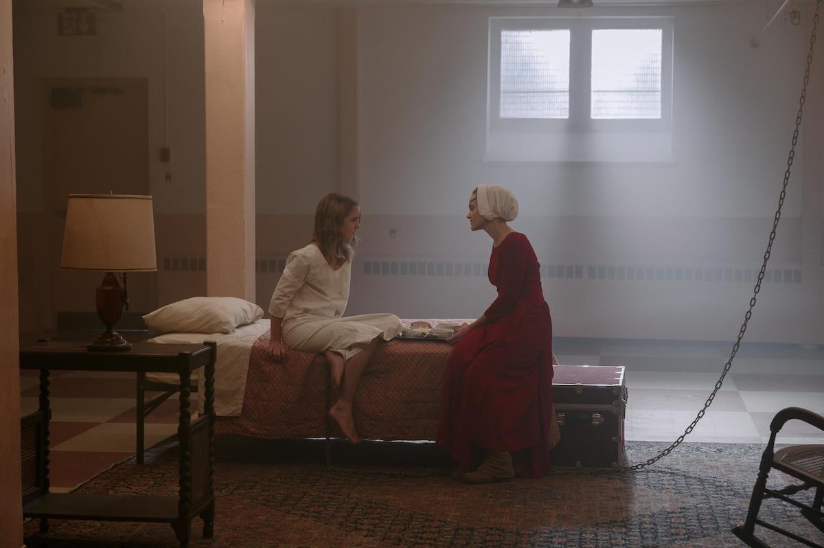 Mckenna Grace as Esther Keyes and Madeline Brewer as Janine in 'The Handmaid's Tale' Season 4. Grace wears a white nightgown and sits on a bed next to Brewer, who wears a red Handmaid dress and white bonnet. They're in a school cafeteria turned into a prison cell. A chain stretches from floor to ceiling next to the bed.