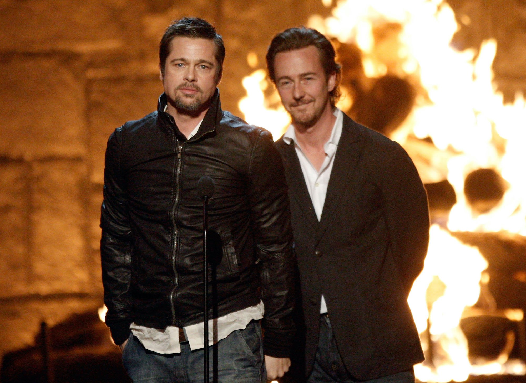 Brad Pitt and Edward Norton behind a microphone, on a stage with a fire in the background