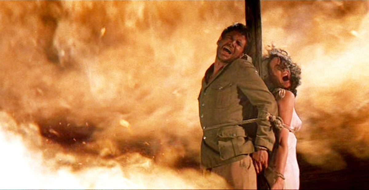 Harrison Ford as Indiana Jones and Karen Allen as Marion Ravenwood in 'Raiders of the Lost Ark'. They're tied to a stake as a fire burns behind them.