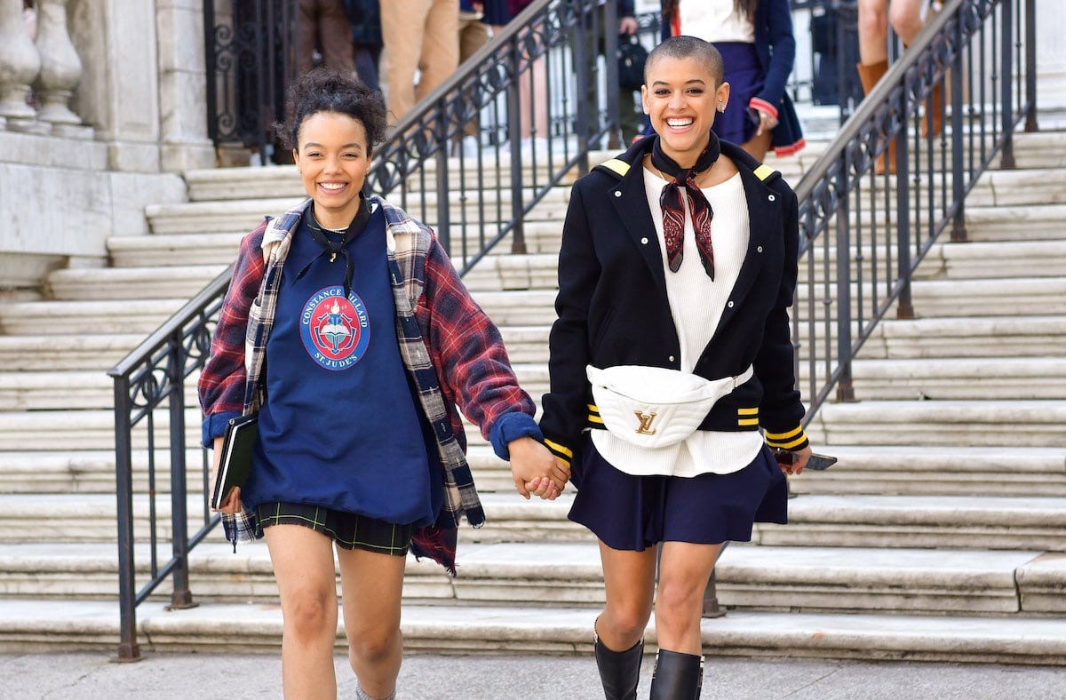 Whitney Peak and Jordan Alexander hold hands on the set of "Gossip Girl" in front of steps of a building.