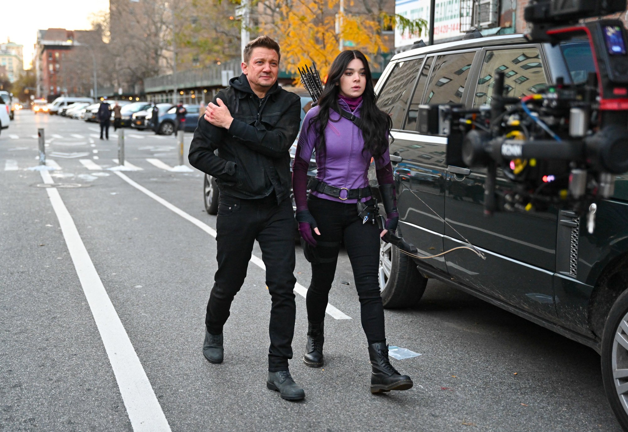 Jeremy Renner and Hailee Steinfeld walking down the street while filming Marvel's Hawkeye series -- She's wearing a black and purple costume and carrying her arrows and he's dressed in all black