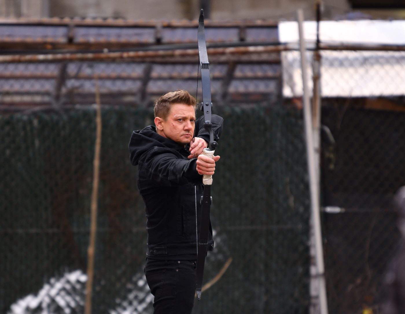 The actor Jeremy Renner stands in front of a blurred background of a high chain link fence with a bow drawn in a pair of black jean pants and a black hooded coat.