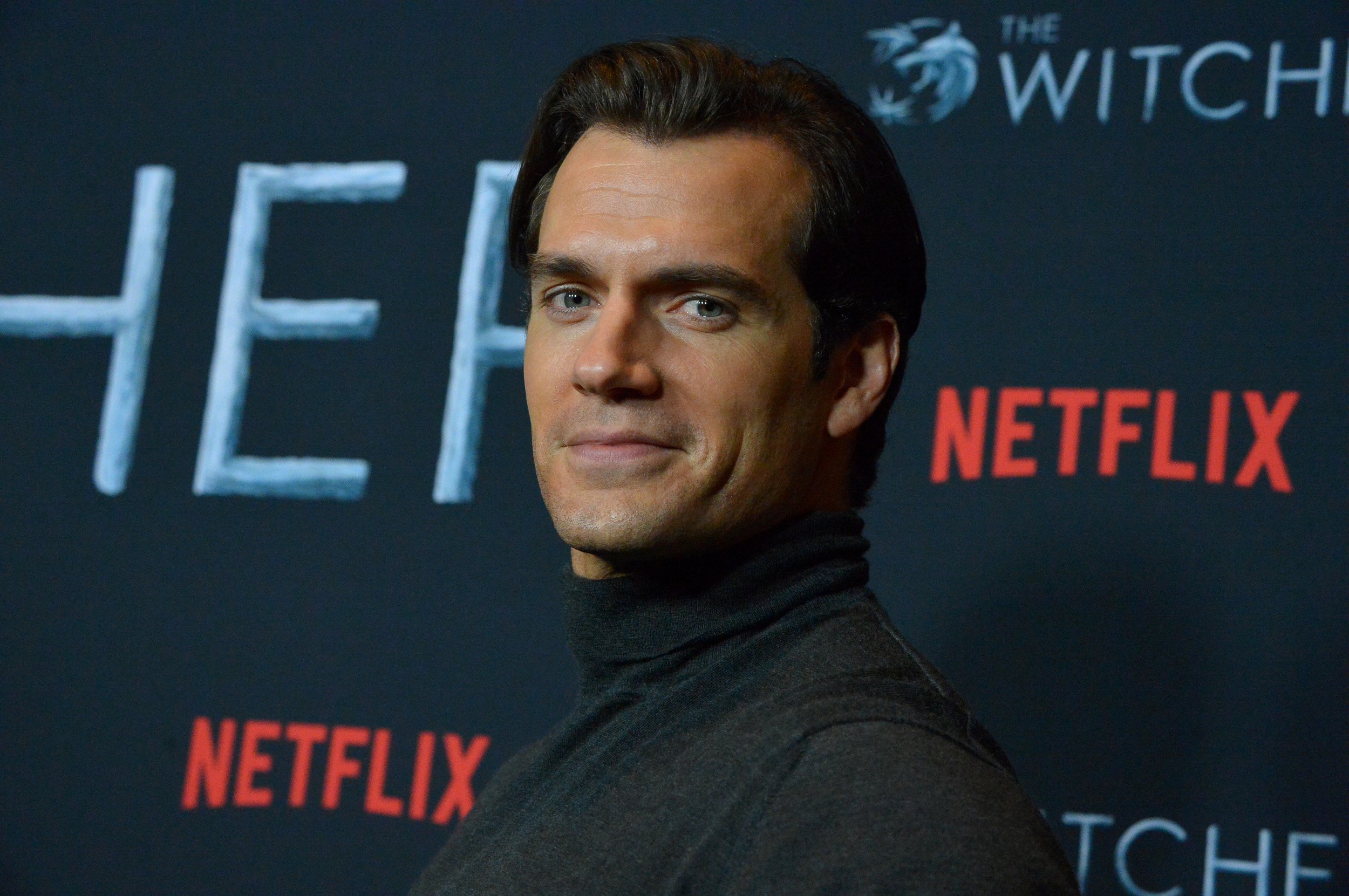 'The Witcher' star Henry Cavill wearing a black shirt and standing in front of a Netflix wall