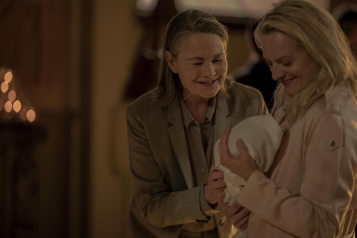 Holly (Cherry Jones) and June (Elisabeth Moss) in 'The Handmaid's Tale' Season 3. Moss wears a pink coat and holds a baby, who's wrapped in a white blanket. Jones wears a beige suit and smiles down at the baby. They're in a church for a baptism. The background is blurred.