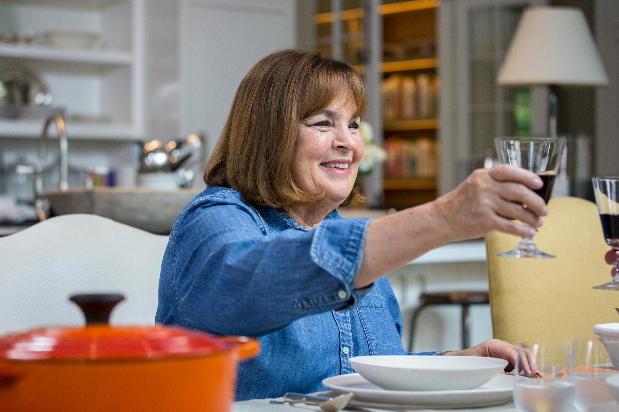 Ina Garten wears a blue shirt while smiling and toasting with a glass of wine