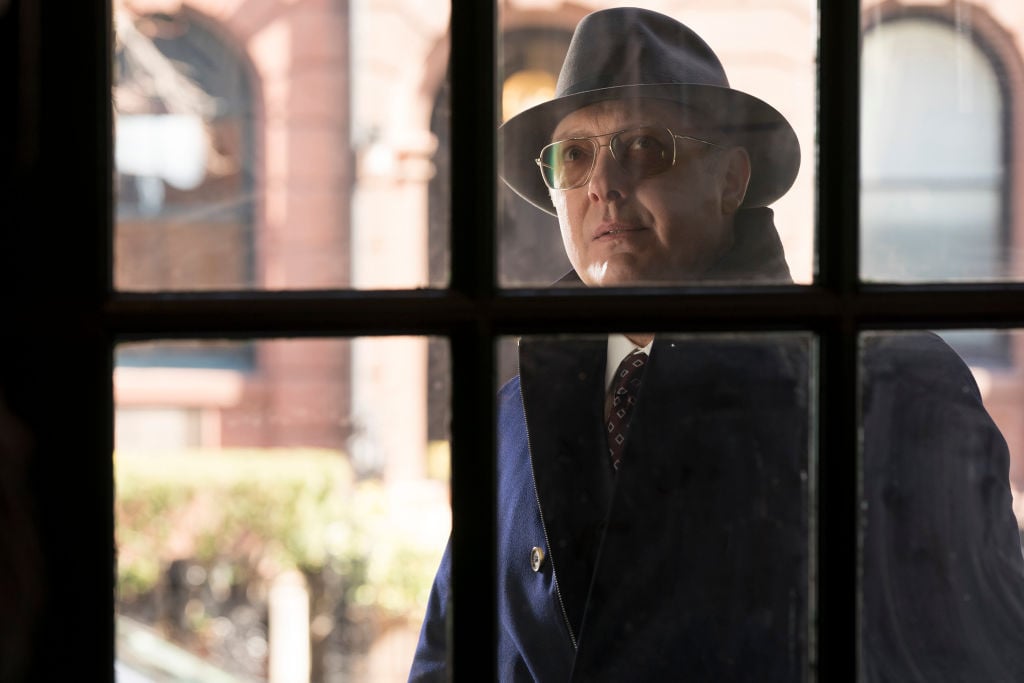 James Spader as Raymond 'Red' Reddington  looks through a window from the outside. He's wearing a trench coat, fedora, and sunglasses.
