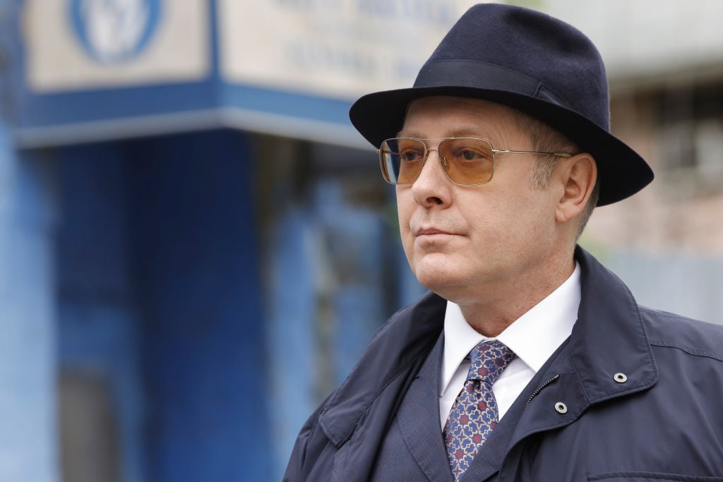 James Spader as Raymond 'Red' Reddington stands outside in his trademark navy fedora, sunglasses, and trenchcoat.