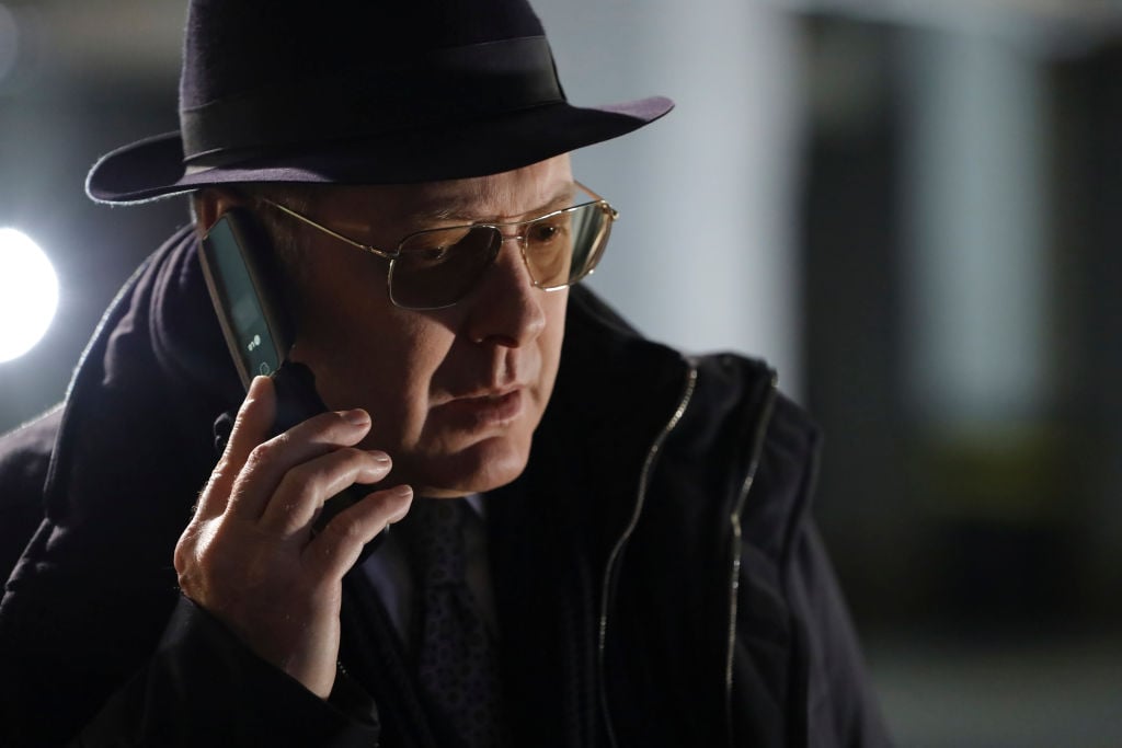 James Spader as Raymond 'Red' Reddington is wearing his classic fedora and glasses while speaking on the phone.