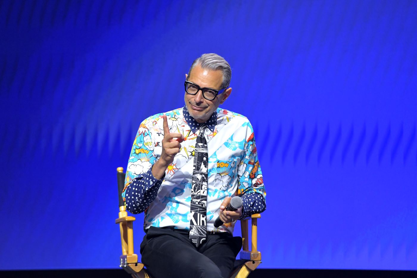 Jeff Goldblum in a very bold-patterned button-up shirt and tie with a dark blue long sleeve shirt underneath against a black background sitting in a director's chair in front of a blue background.
