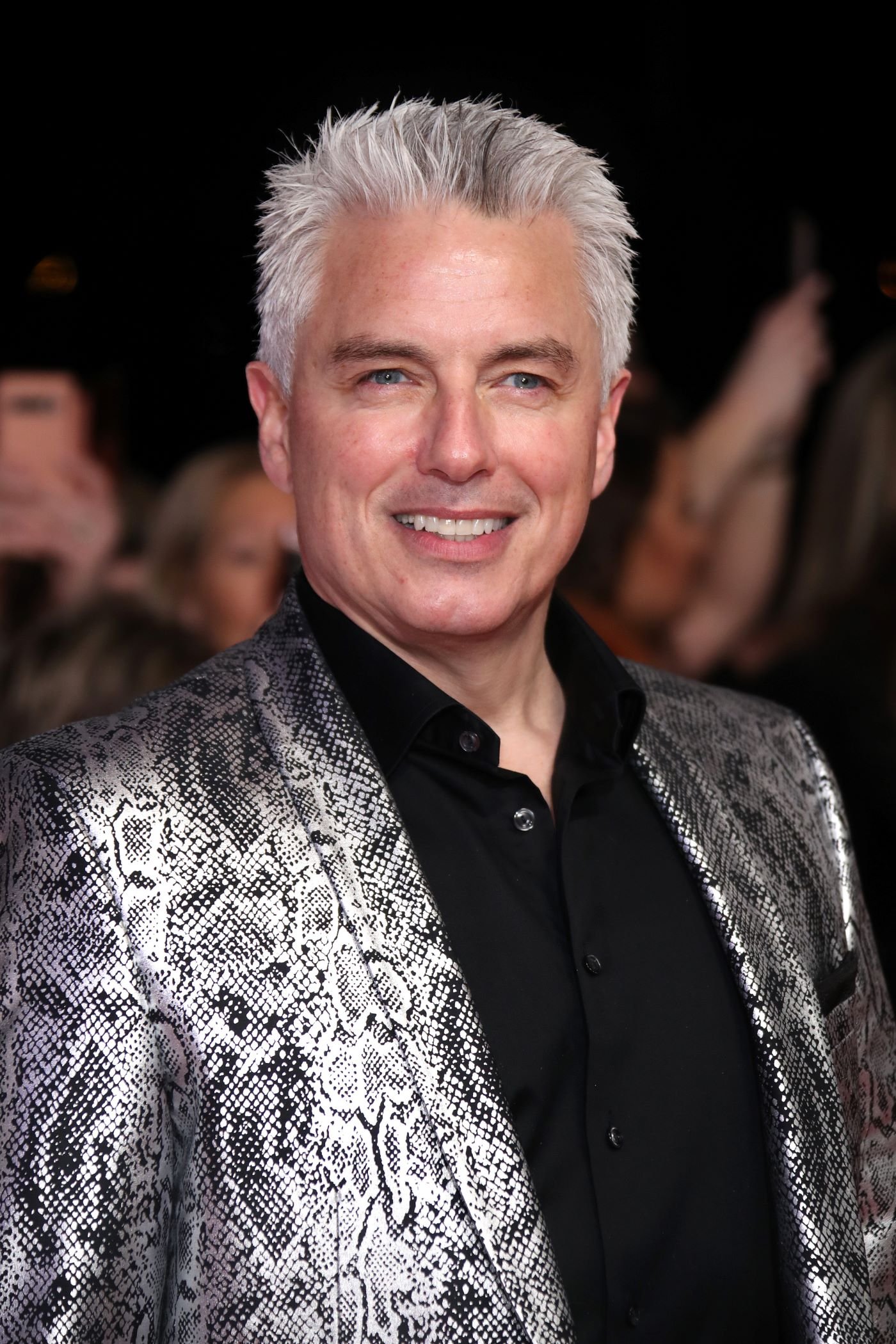 John Borrowman stands in front of a blurred black background with people in it wearing a black button-up shirt and a silver and black metallic snake print suit jacket.
