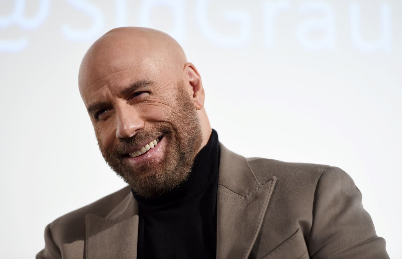 John Travolta stands in front of a white background wearing a black turtle neck and a light brown suit jacket.