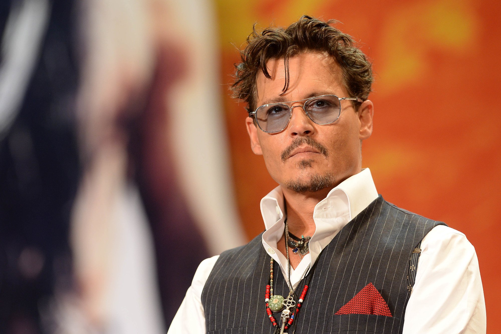 Johnny Depp wearing glasses, a white collared shirt, and a striped black and white vest. His hair is gelled back and one piece hangs in front of his face. He's wearing a blank expression.